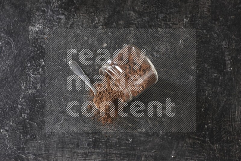 A flipped glass jar and metal spoon full of cloves powder with cloves spread on a textured black flooring