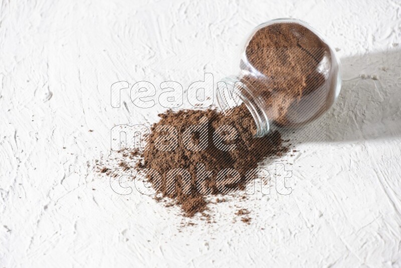 A flipped glass spice jar full of cloves powder and powder came out of it on textured white flooring