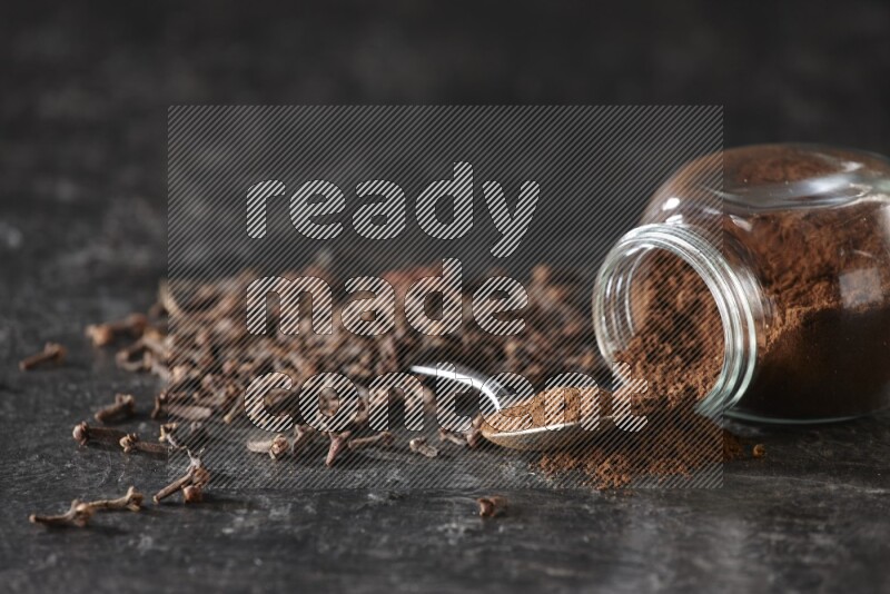 A flipped glass spice jar and a metal spoon full of cloves powder with cloves spread on textured black flooring