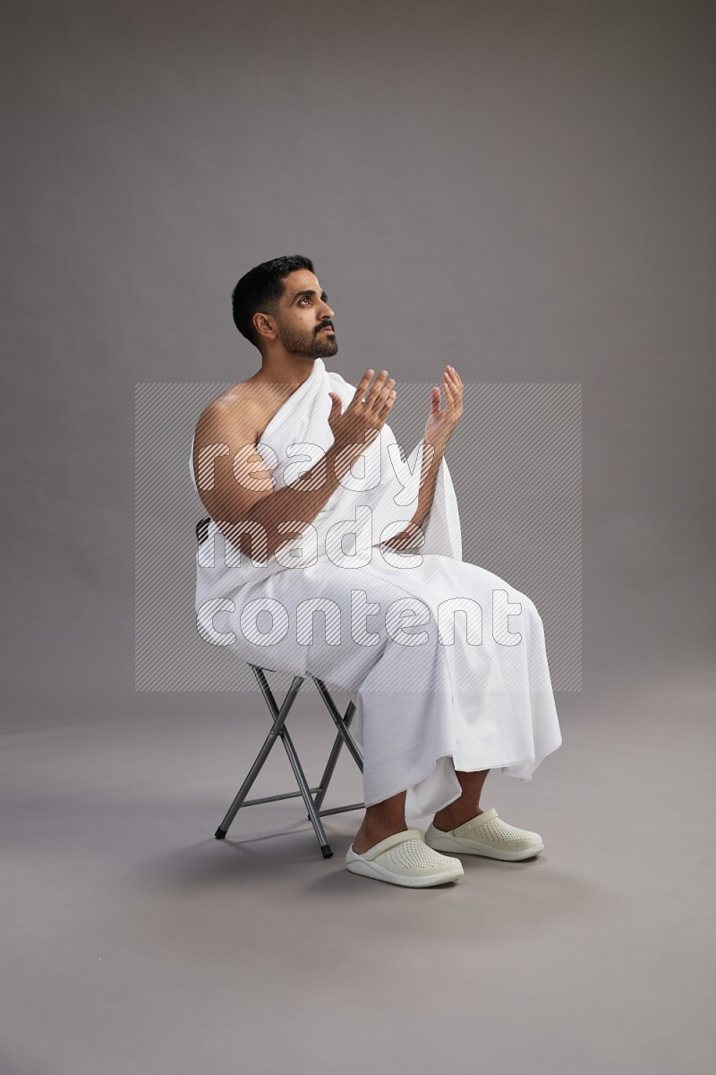 A man wearing Ehram sitting on chair performing dua'a on gray background
