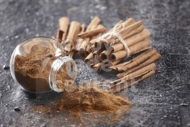 Herbal glass jar full of cinnamon powder flipped with cinnamon sticks stacked and bounded on a textured black background