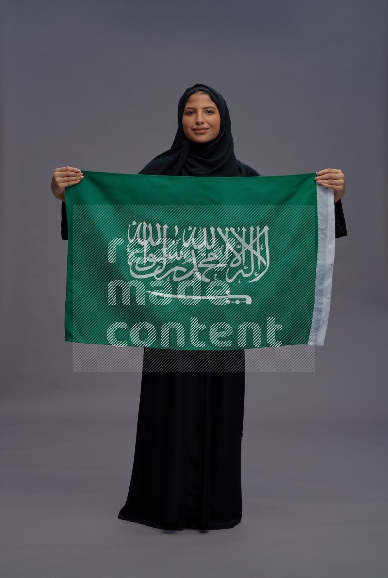 Saudi woman wearing Abaya standing holding Saudi flag on gray background