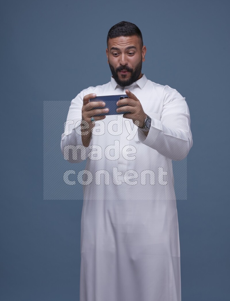 A man Playing a Game on his Smartphone  on Blue Background wearing Saudi Thob