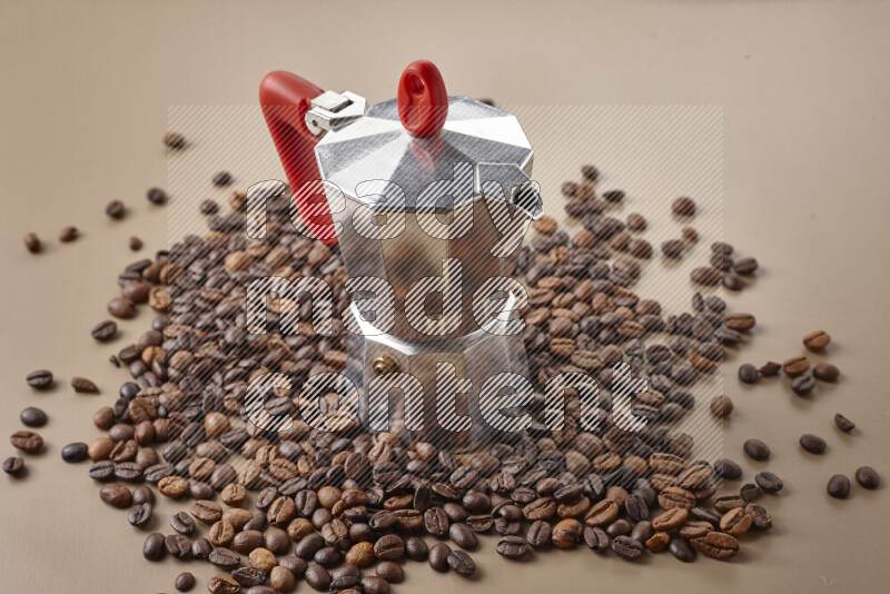 A moka pot with red handle surrounded by roasted coffee beans on beige background