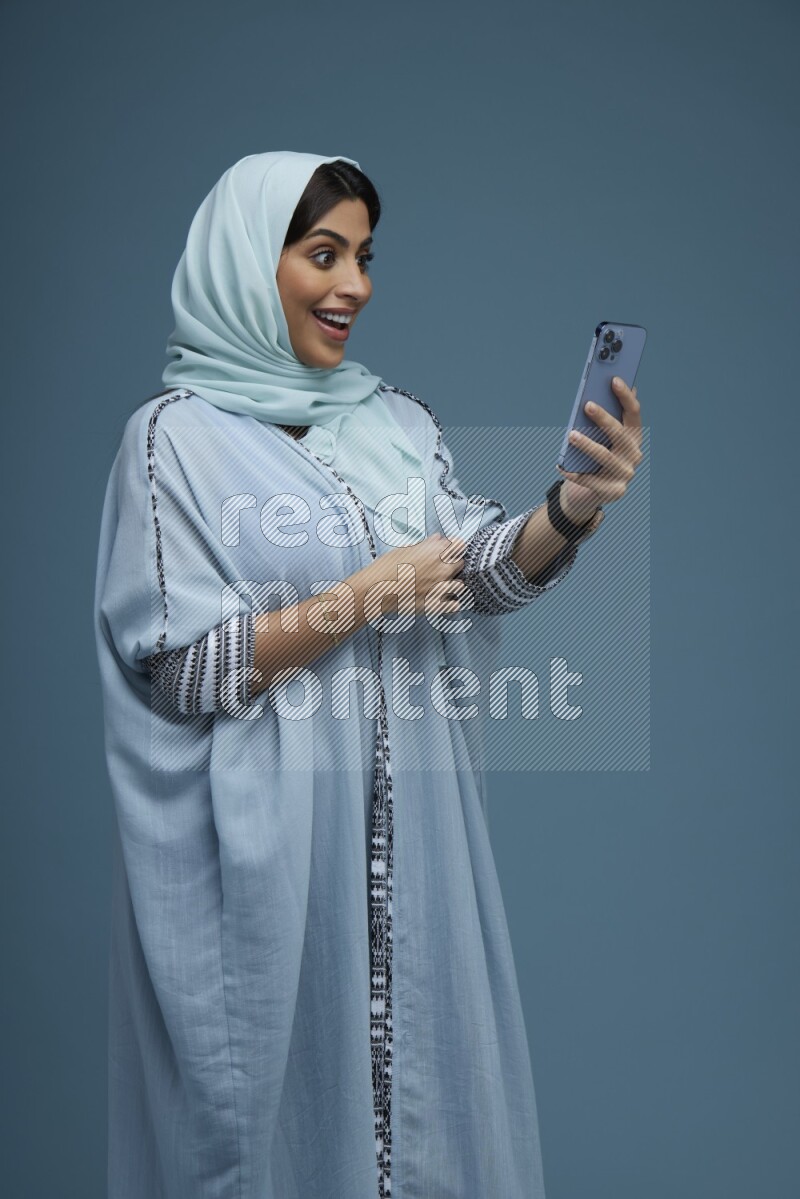 A Saudi woman posing with her phone in a blue background wearing a blue Abaya with hijab