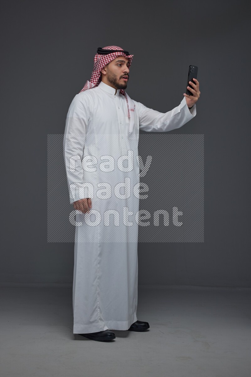 A Saudi man wearing Thobe and Shamgh taking selfie with his phone eye level on a gray background
