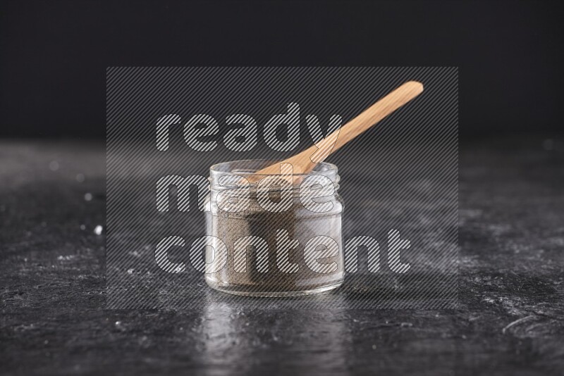 A glass jar full of black pepper powder and a wooden spoon on a textured black flooring