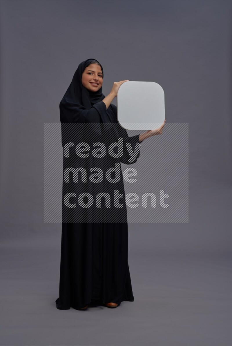 Saudi woman wearing Abaya standing holding social media sign on gray background