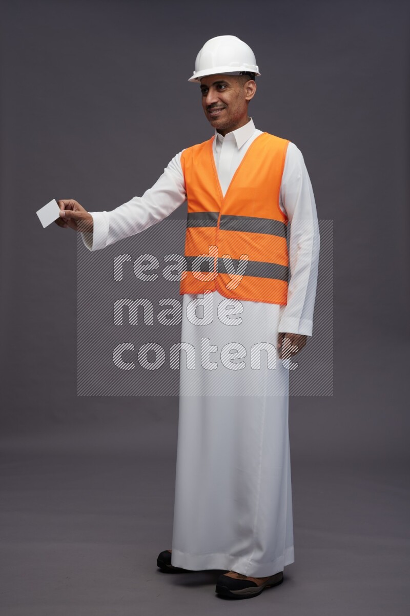 Saudi man wearing thob with engineer vest standing holding ATM card on gray background