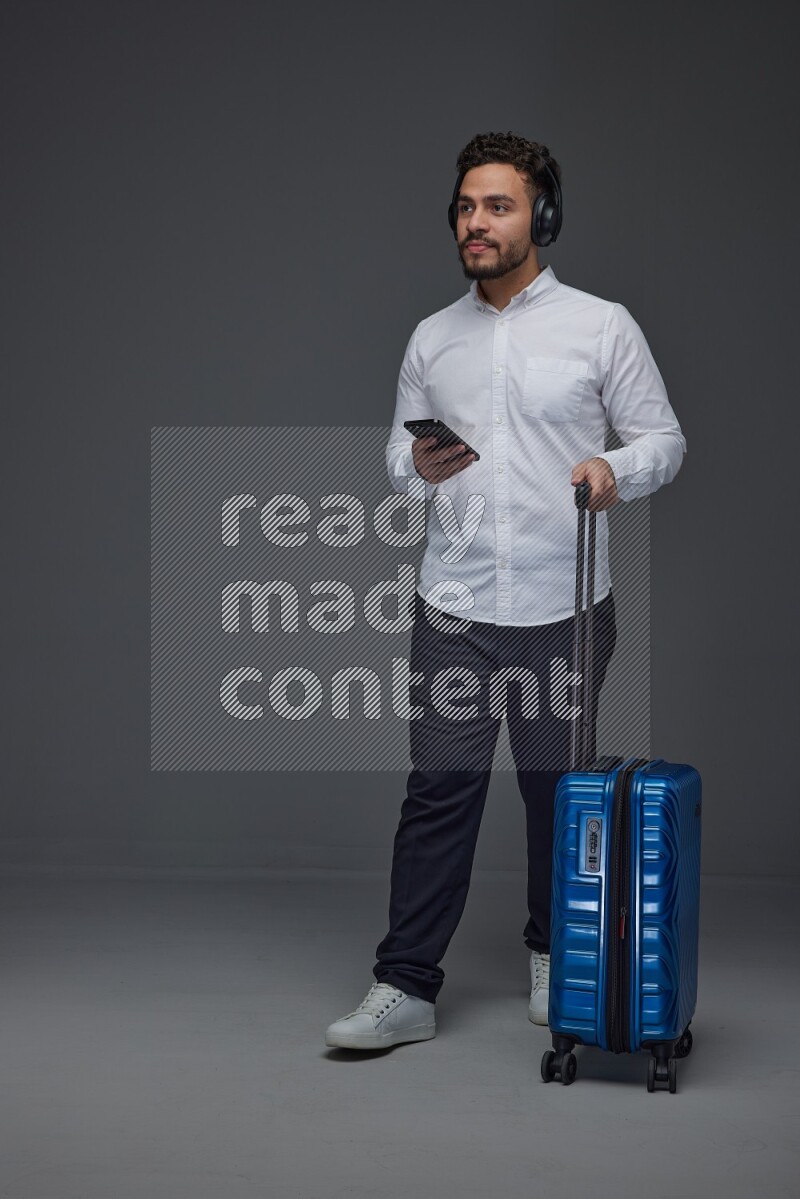 A man wearing smart casual using his phone while wearing headphone and holding luggage eye level on a gray background