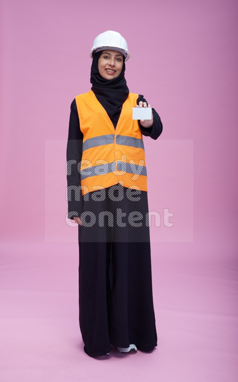 Saudi woman wearing Abaya with engineer vest and helmet standing holding ATM card on pink background
