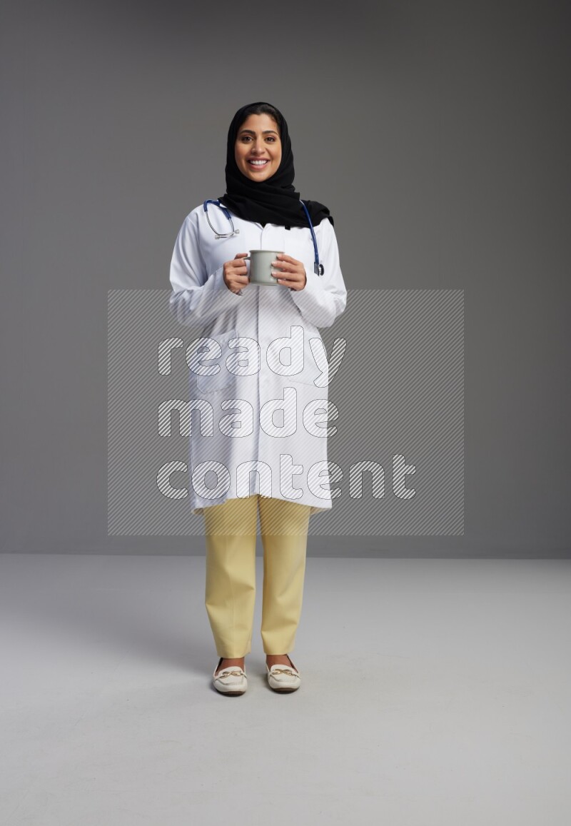 Saudi woman wearing lab coat with stethoscope standing holding mug on Gray background