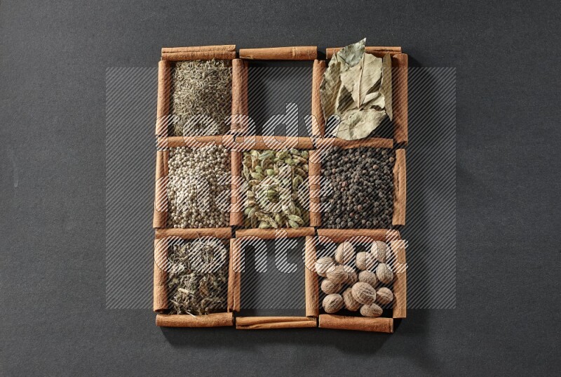 9 مربعات من أعواد القرفة ممتلئة بالهيل في المنتصف محاطة بجوزة الطيب والفلفل الأسود وورق الغار والبهارات والكمون والفلفل الأبيض والريحان المجفف والملح على أرضية سوداء