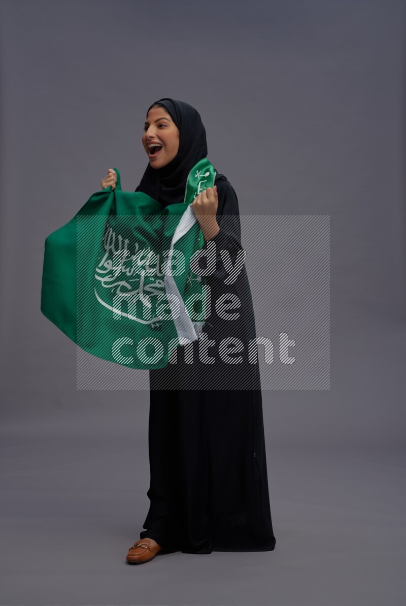 Saudi woman wearing Abaya standing holding Saudi flag on gray background