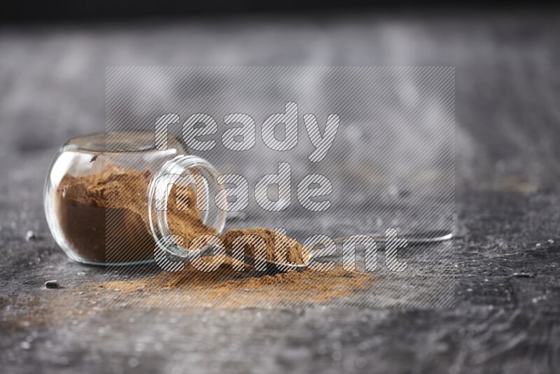 Herbal glass jar full of cinnamon powder flipped and a metal spoon on textured black background