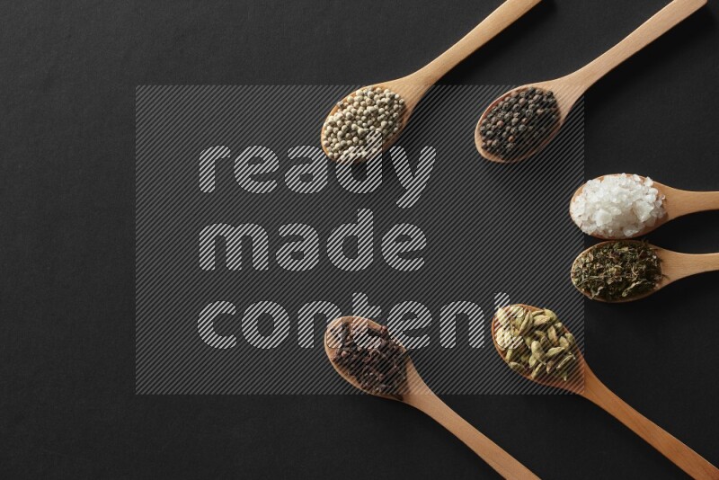 الفلفل الأبيض والقرنفل والهيل والملح والفلفل الأسود وريحان مجفف في معالق خشبية علي خلفية سوداء