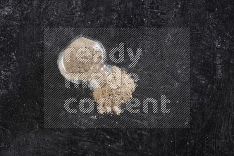 A glass spice jar full of garlic powder flipped over with the powder came out on a textured black flooring