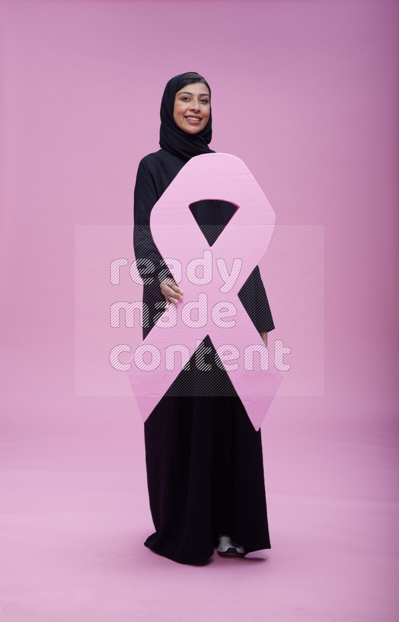 Saudi woman wearing pink ribbon on abaya standing holding awareness ribbon on plain pink background