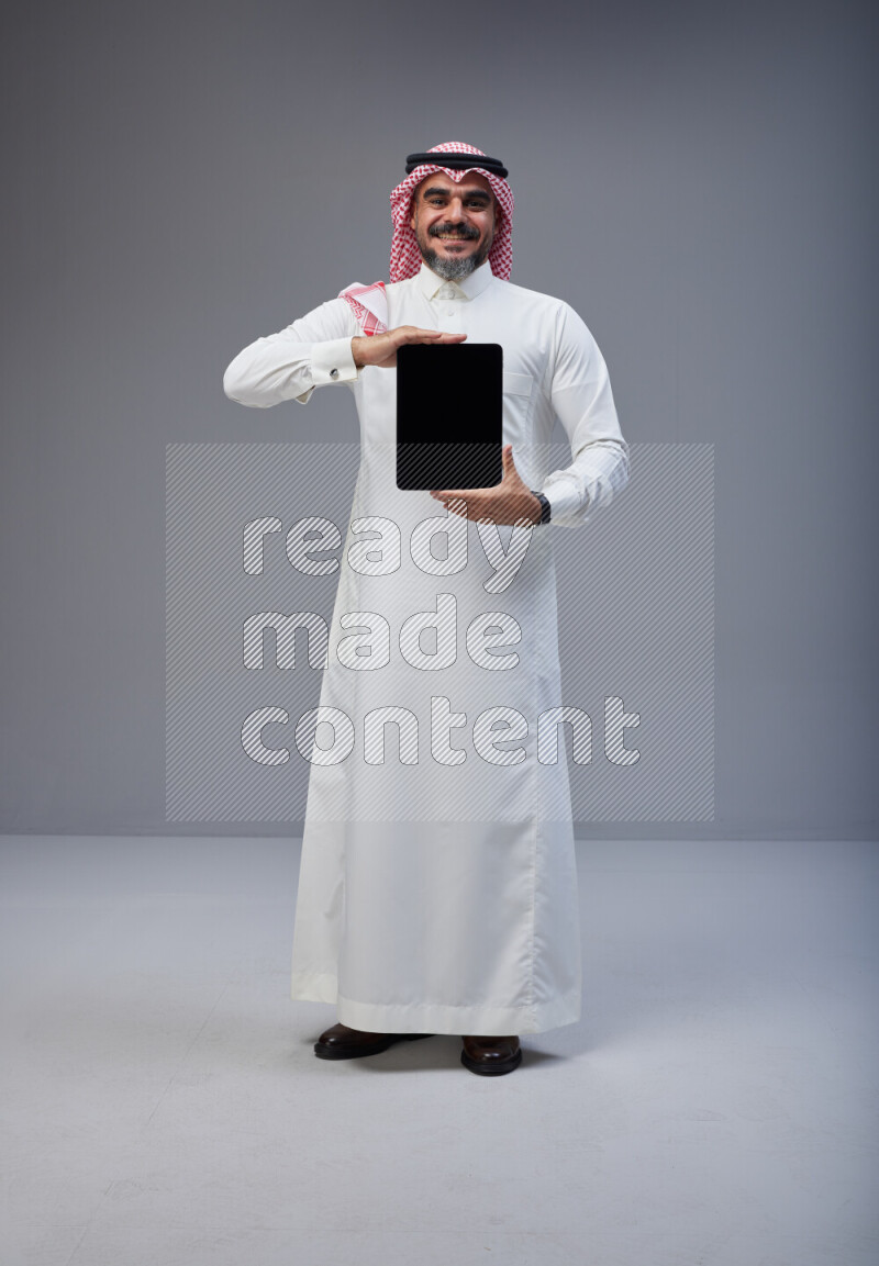 Saudi man Wearing Thob and red Shomag standing showing tablet to camera on Gray background