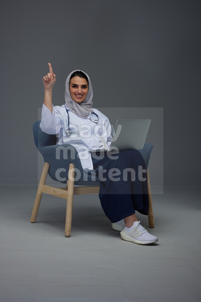 A doctor wearing a light gray head scarf setting on a dark grey chair and using a phone eye level on a grey background