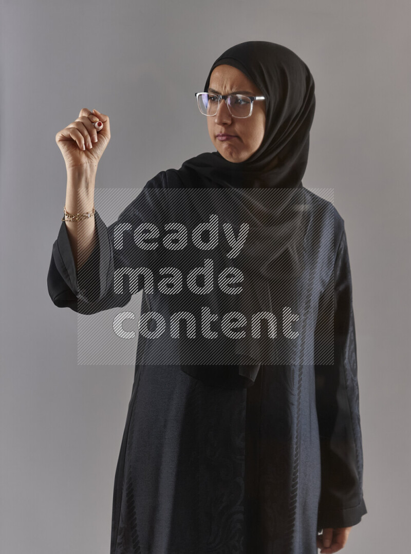 Woman in black abaya, hijab and glasses holding a marker pen to write on a transparent board with different reactions and expressions on grey background