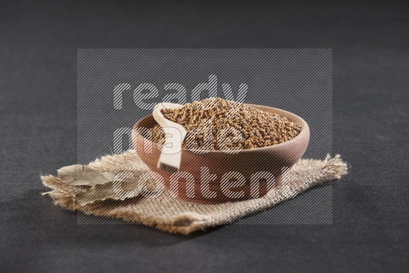 A wooden bowl and spoon full of mustard seeds on a piece of burlap on a black flooring