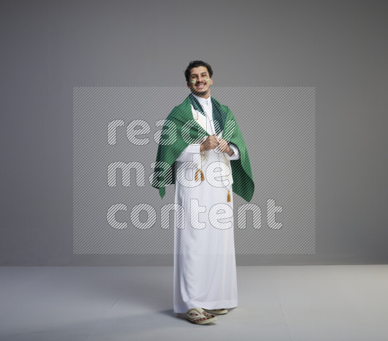 A Saudi man standing wearing thob and saudi flag scarf with face painting wrapping big Saudi flag on gray background