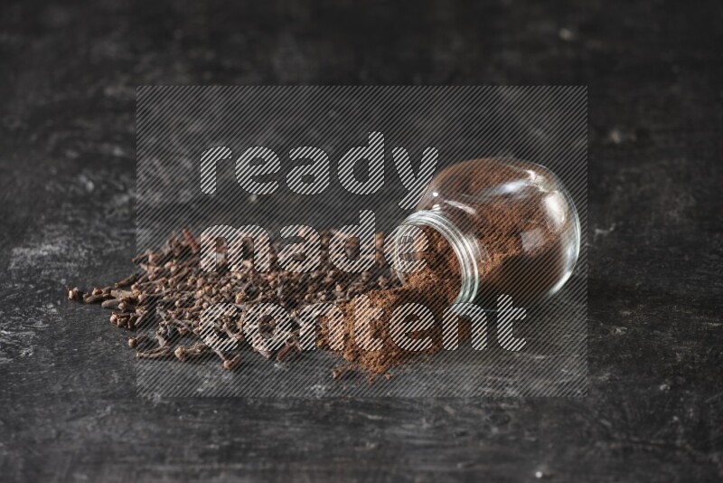 وعاء زجاجي للبهارات مقلوب ممتلئ ببودرة القرنفل والبودرة سقطت منه مع حبوب القرنفل المتناثرة على أرضية سوداء