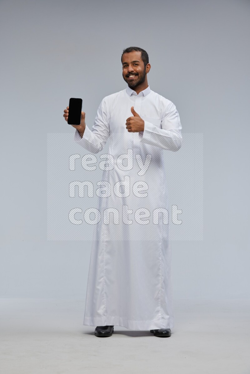 Saudi man wearing Thob standing showing phone to camera on Gray background