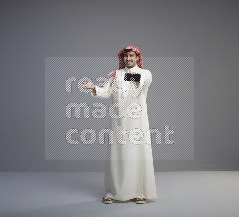 A Saudi man standing wearing thob and red shomag showing phone to camera on gray background