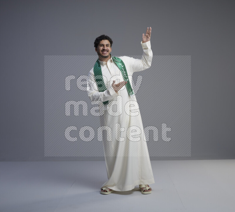 A Saudi man standing wearing thob and Saudi flag scarf on gray background