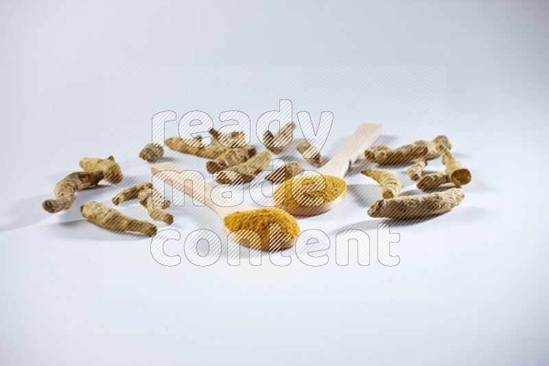 ملعقتان خشبيتان ممتلئتان ببودرة الكركم مع أصابع الكركم المجففة بجانبهما على خلفية بيضاء