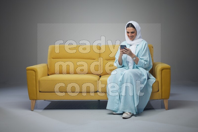 A Saudi woman wearing a light blue Abaya and white head scarf sitting on a yellow sofa and texting on her phone on a grey background