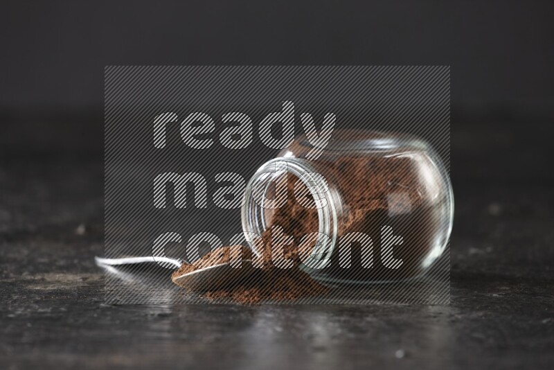 A flipped glass spice jar and a metal spoon full of cloves powder on textured black flooring