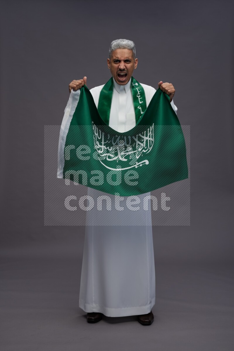 Saudi man wearing thob standing holding Saudi flag on gray background