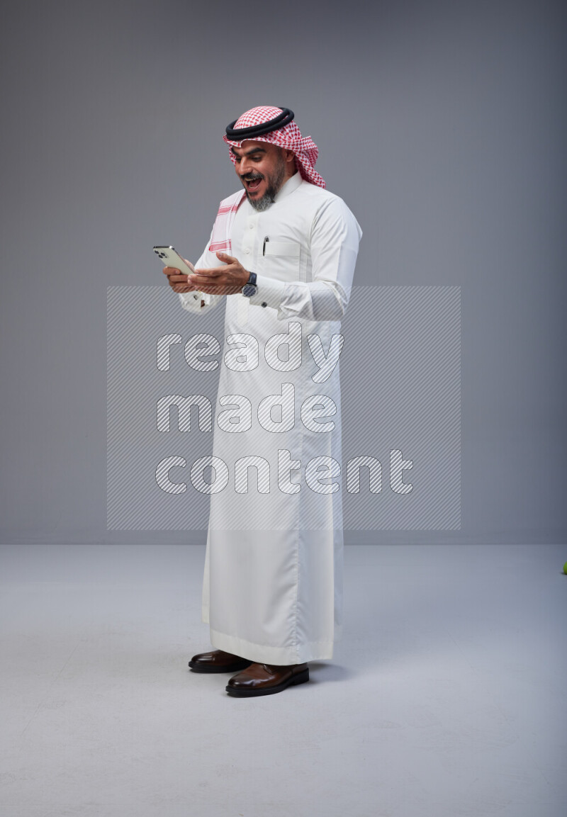 Saudi man Wearing Thob and red Shomag standing texting on phone on Gray background