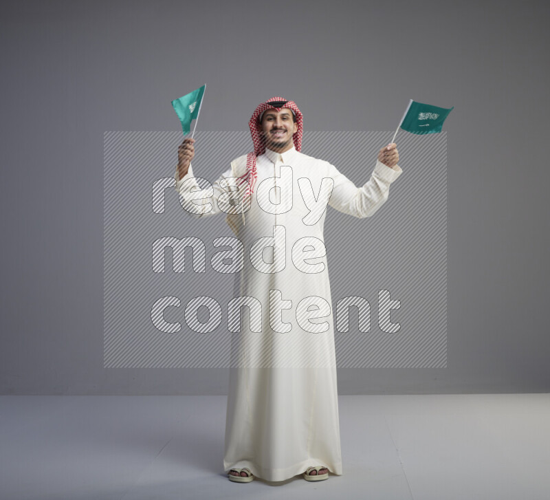 A Saudi man standing wearing thob and red shomag raising small Saudi flag on gray background