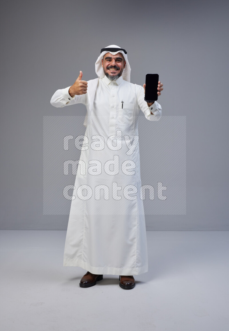 Saudi man Wearing Thob and white Shomag standing showing phone to camera on Gray background