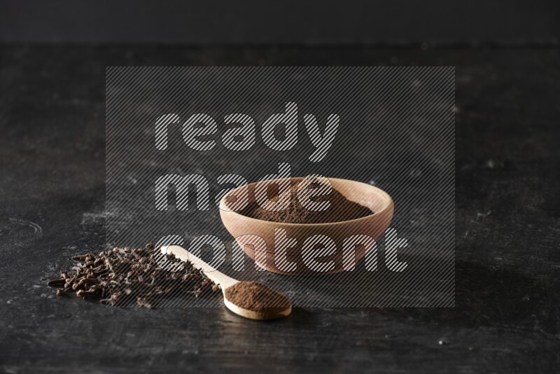 A wooden bowl and a wooden spoon full of cloves powder with spreaded cloves on a textured black flooring