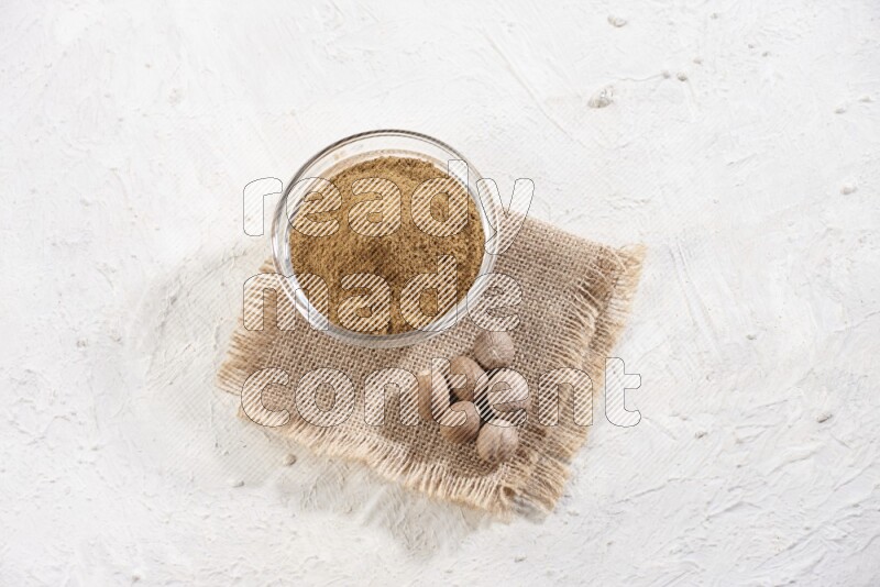A glass bowl full of nutmeg powder with whole seeds beside it on burlap fabric on a textured white flooring
