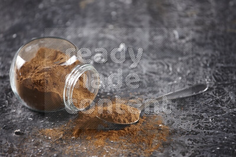 Herbal glass jar full of cinnamon powder flipped and a metal spoon on textured black background