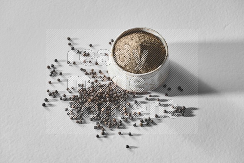 A white ceramic bowl full of black pepper powder and black pepper beads spread on white flooring
