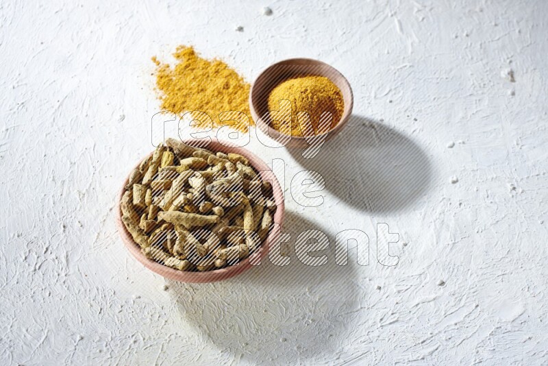 2 wooden bowls, one full of turmeric powder and the other full of dried turmeric whole fingers on a textured white flooring