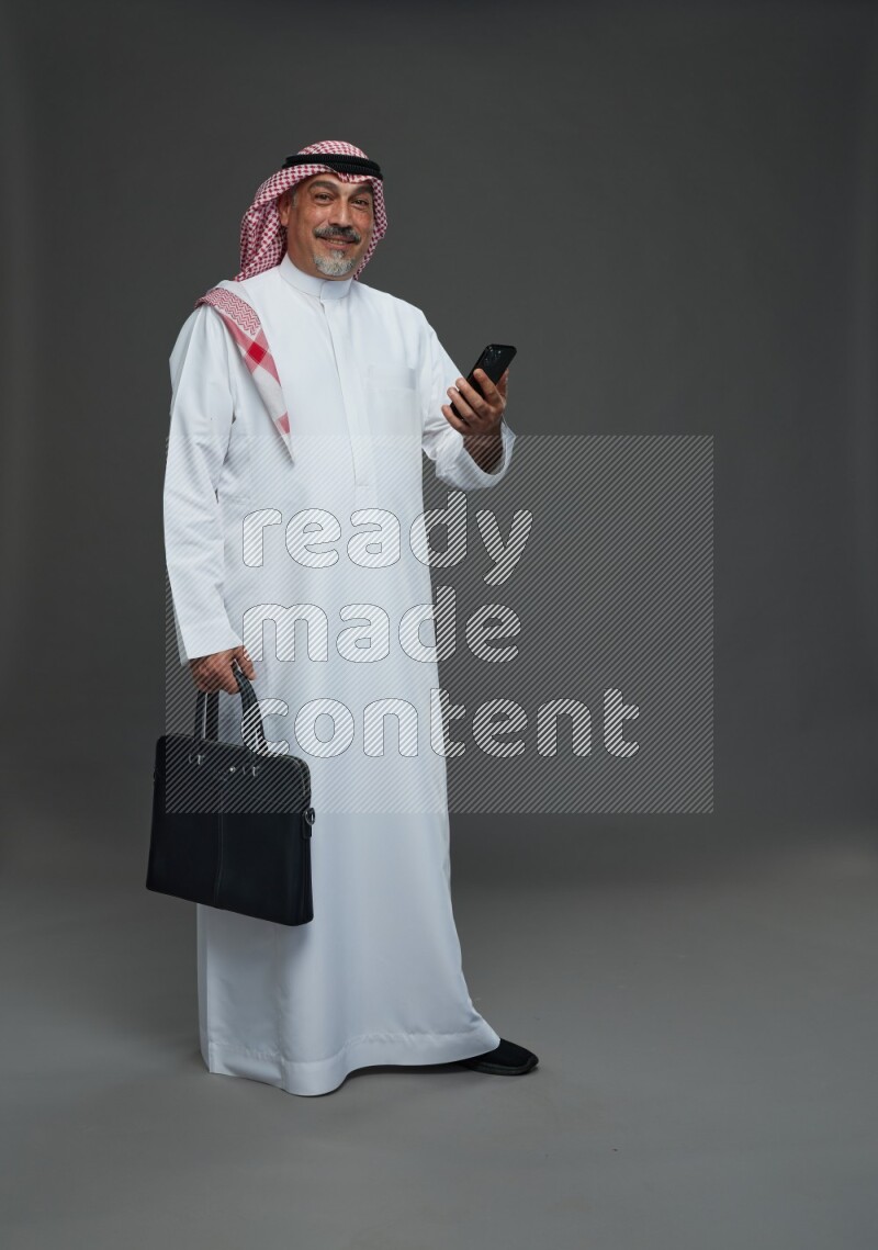 Saudi man with shomag Standing holding bag and phone on gray background
