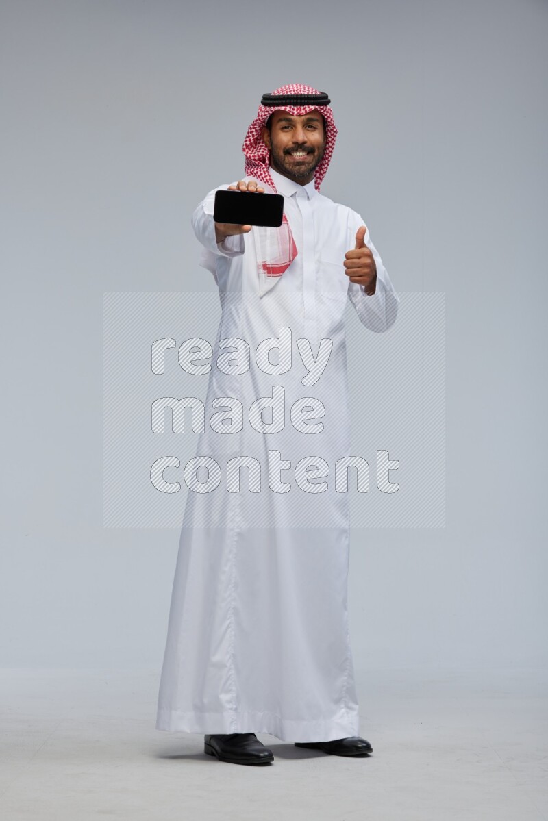 Saudi man Wearing Thob and shomag standing showing phone to camera on Gray background
