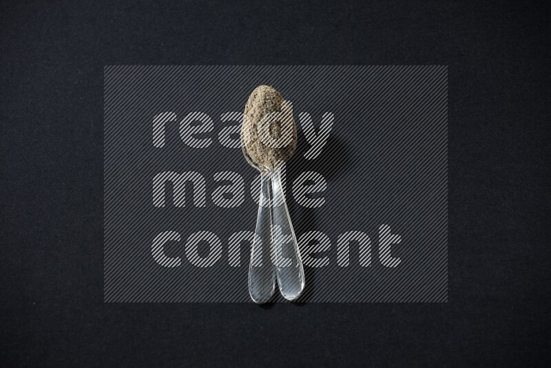 2 Metal spoons full of cardamom powder on black flooring