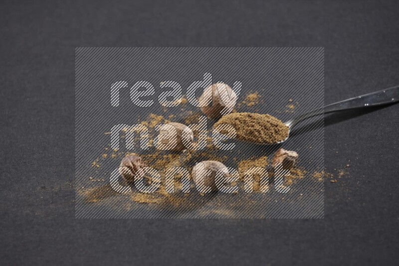 A metal spoon full of nutmeg powder with nutmeg powder and whole seeds beside it on a black flooring