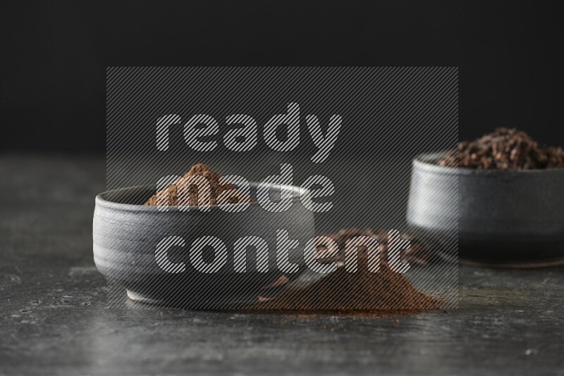 2 Black pottery bowls full of cloves and the other full of cloves powder on textured black flooring
