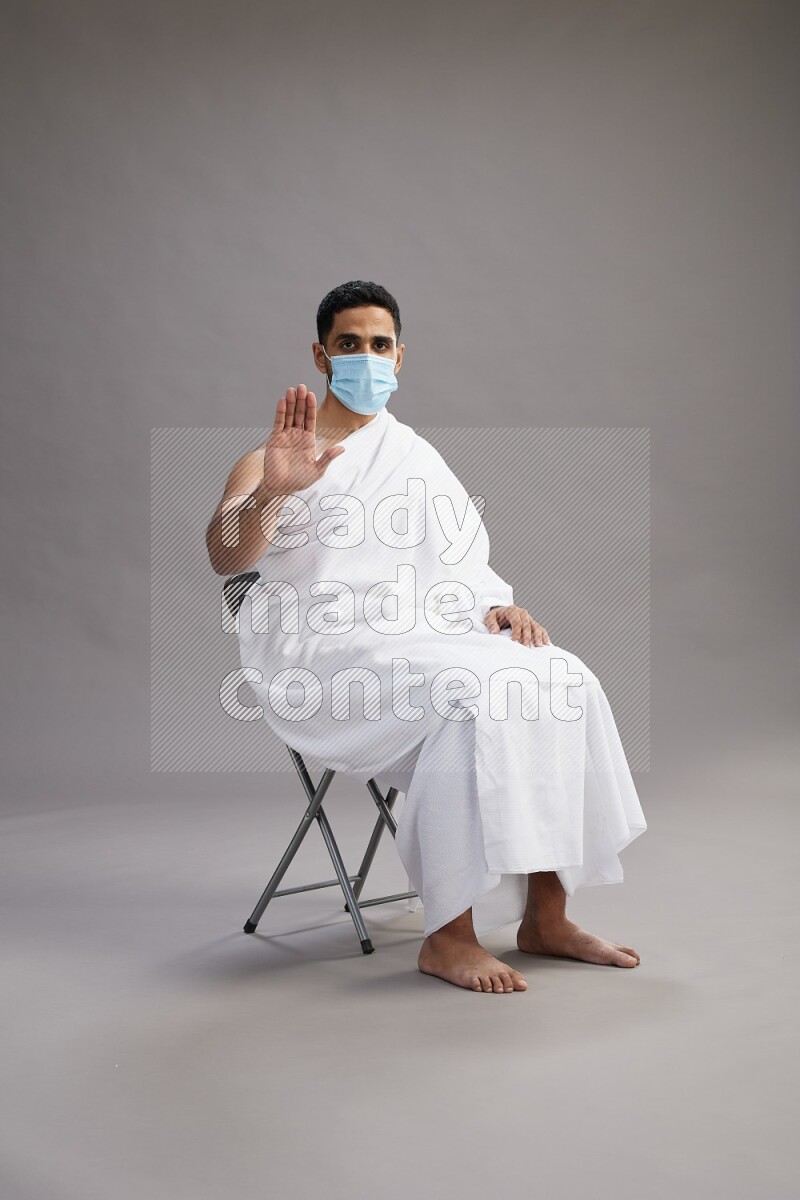 A man wearing Ehram with face mask sitting on chair Interacting with the camera on gray background