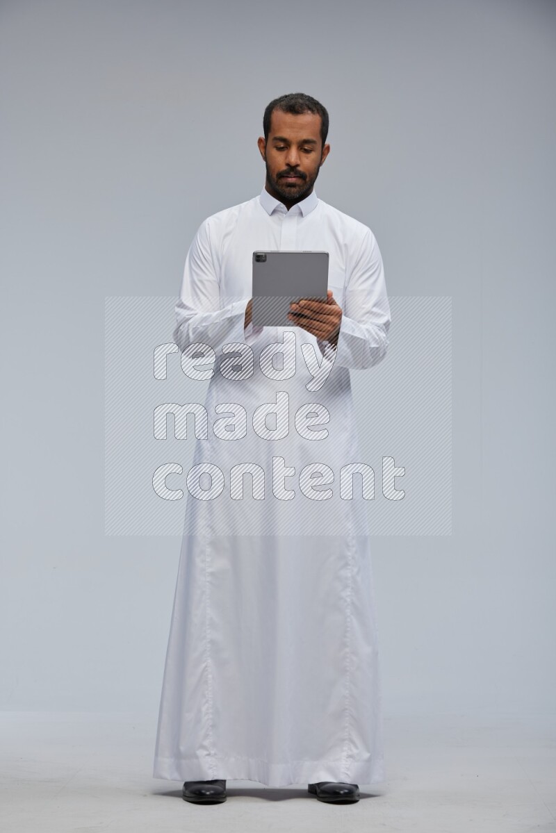 Saudi man wearing Thob standing working on tablet on Gray background